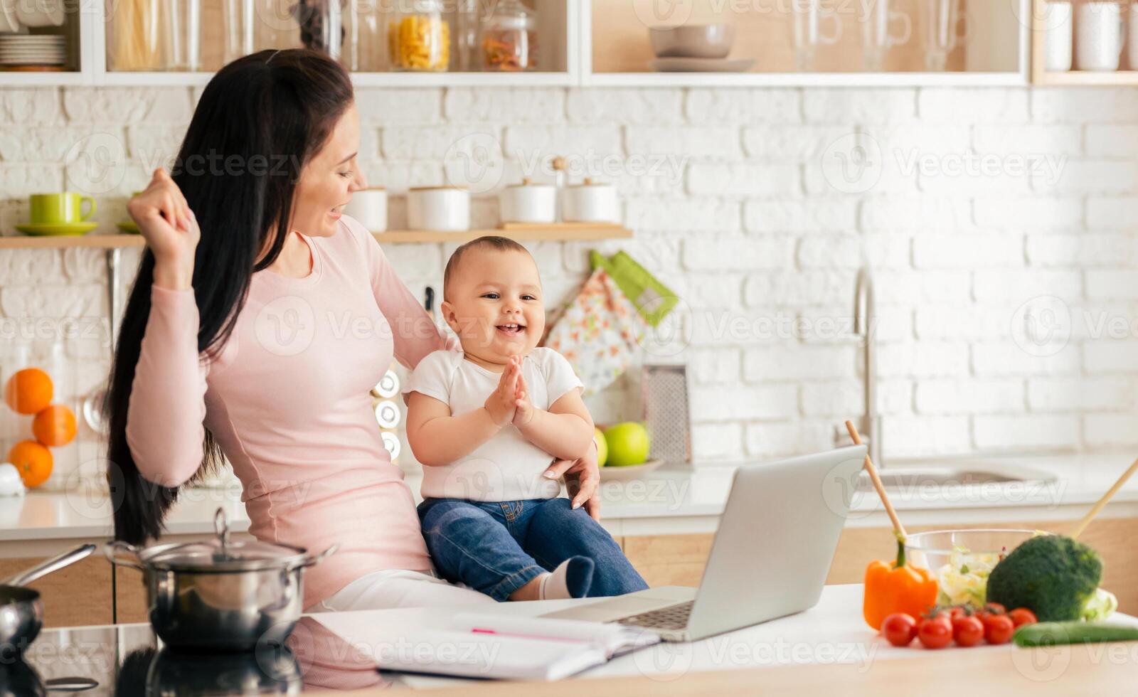 A woman is holding a baby in her arms while looking attentively at a laptop screen. She appears focused and engaged in the digital content displayed on the device. photo