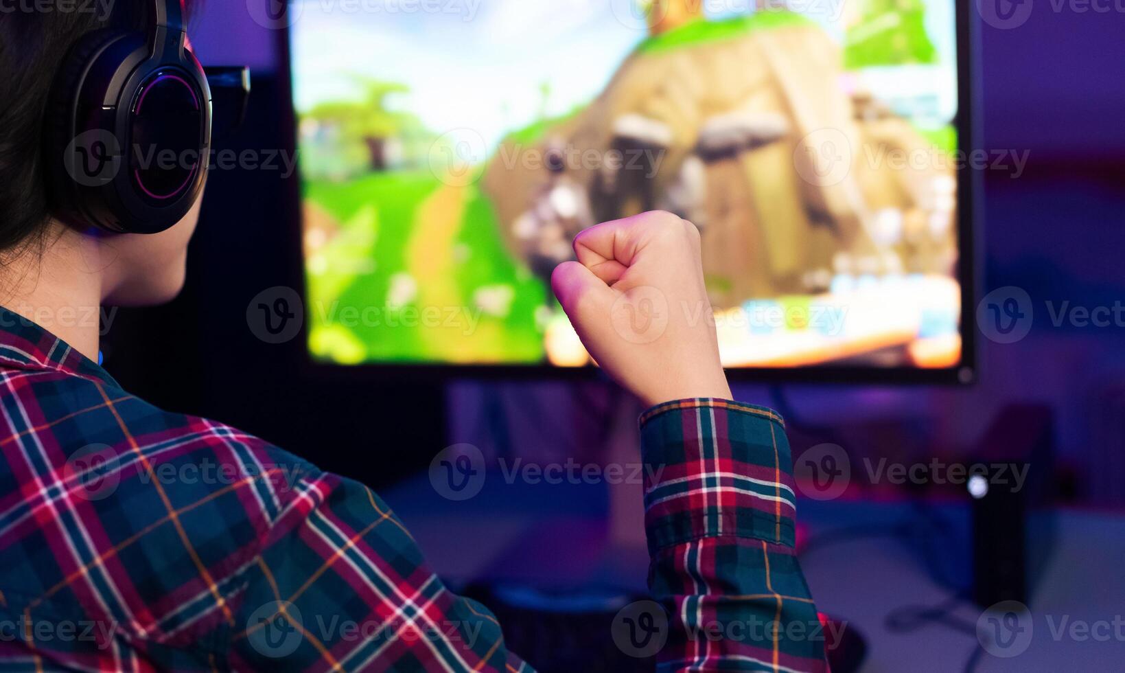 A gamer in a plaid shirt celebrates a victory while playing a game on their computer. They are wearing headphones and have their fist raised in the air. photo