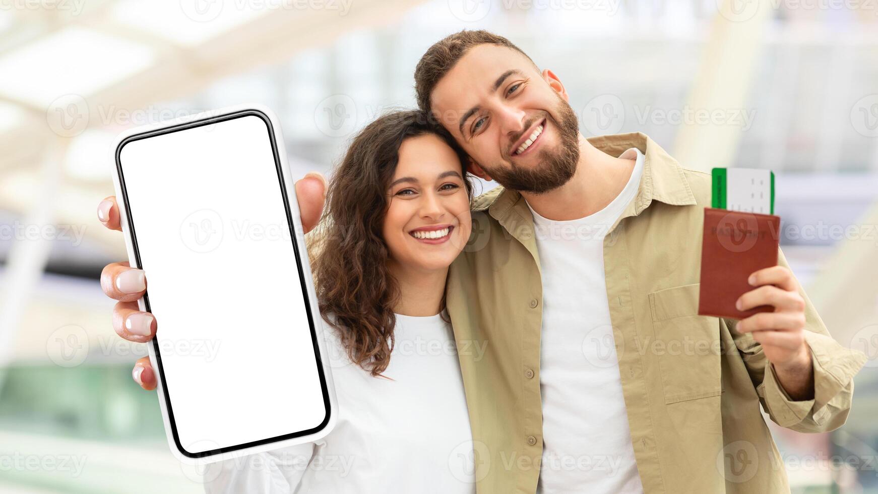 A cheerful man and woman are shown close together holding up a smartphone with a blank screen and a passport. They seem to be in a well-lit airport, possibly ready for travel photo
