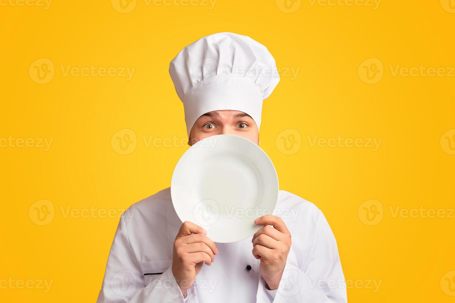 Funny Chef Guy Peeking Out Of Plate Looking At Camera Standing In Studio Over Yellow Background. Panorama photo