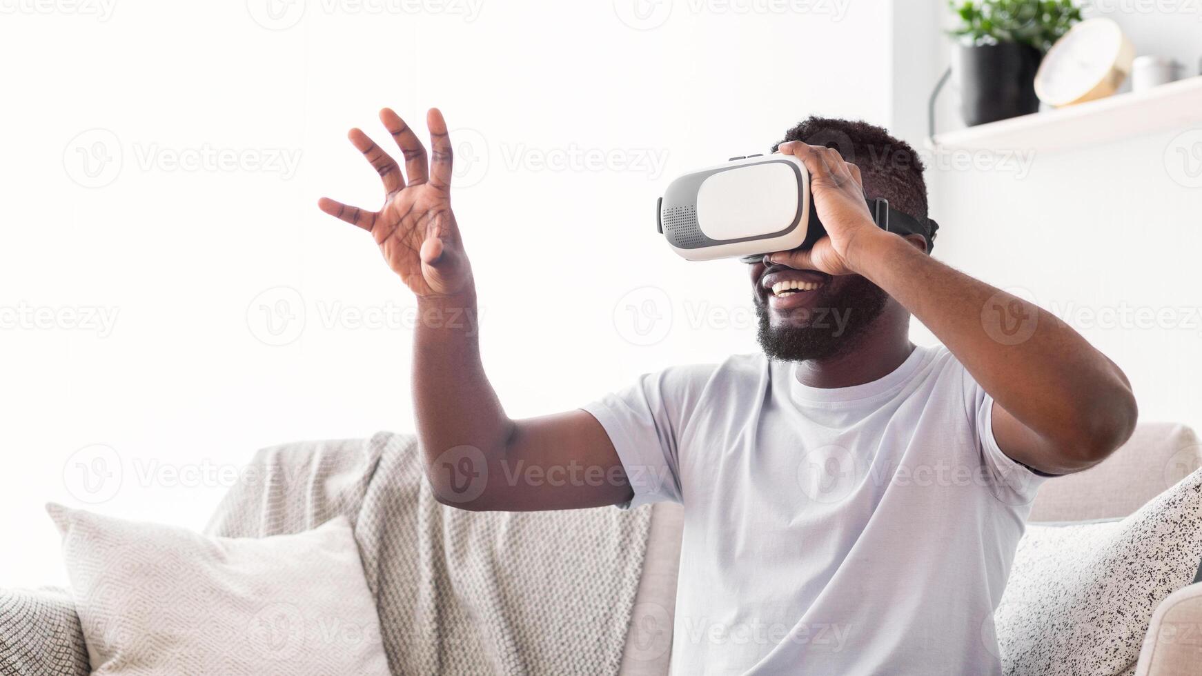 Black man wearing a white t-shirt sits on a light-colored couch and smiles while wearing a virtual reality headset. He raises his right arm as if reaching for something in the virtual world. photo