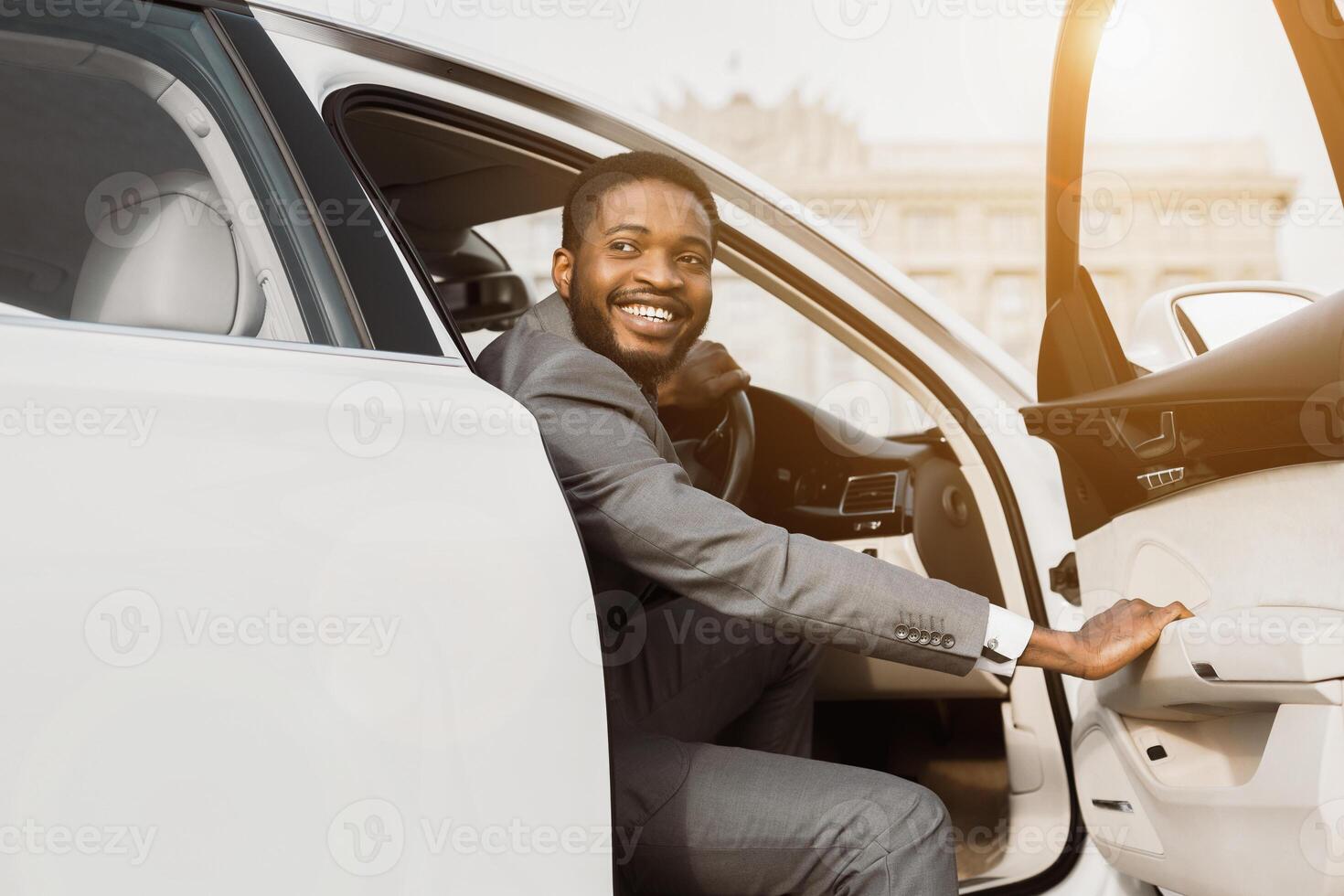 A smiling black businessman in a gray suit exits a white car on a sunny day. The car is parked on a street in an urban setting. He is likely heading to a meeting or appointment. photo