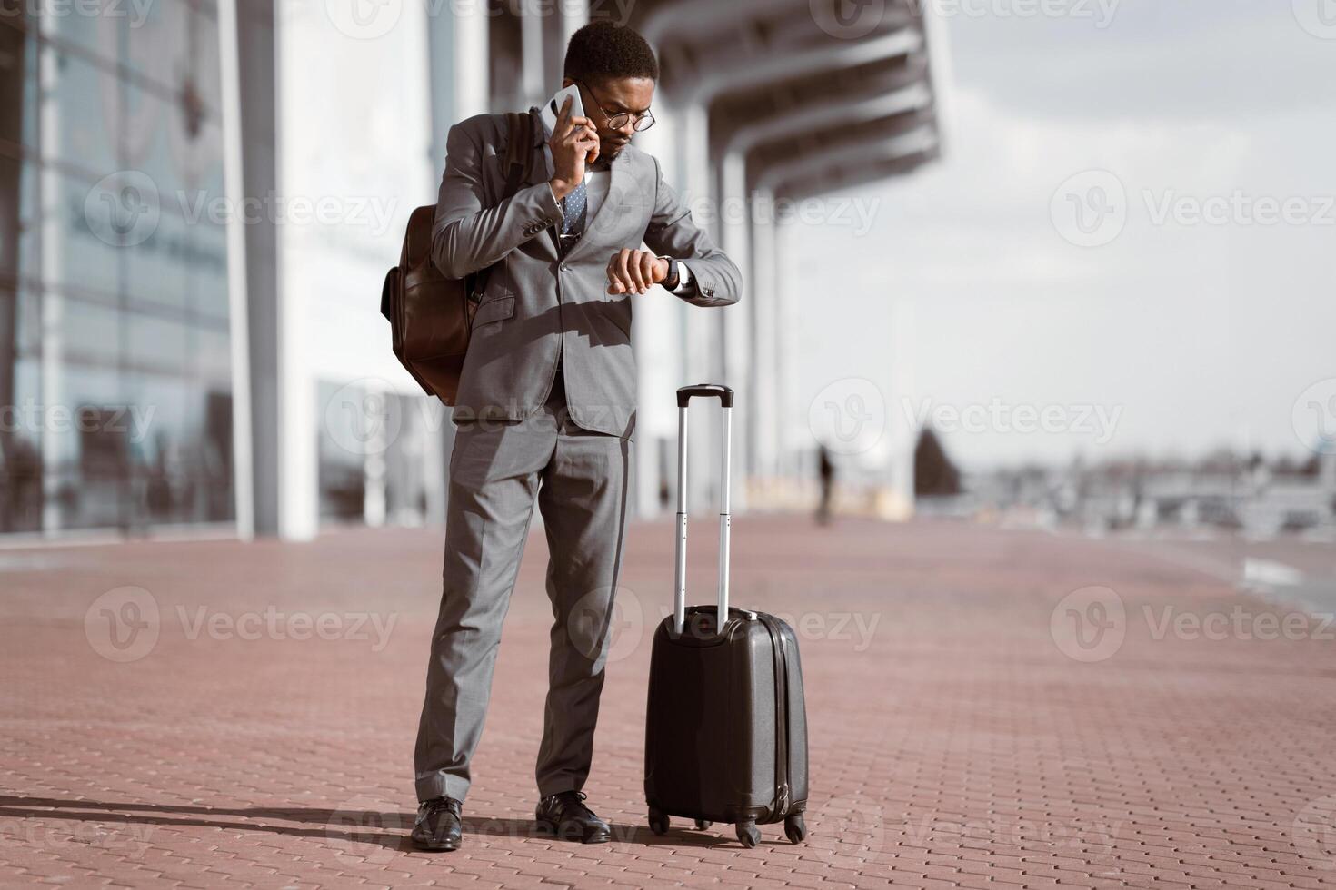 African American businessman on the phone with rolling suitcase outside a modern building, checking time, preparing for a business trip. photo