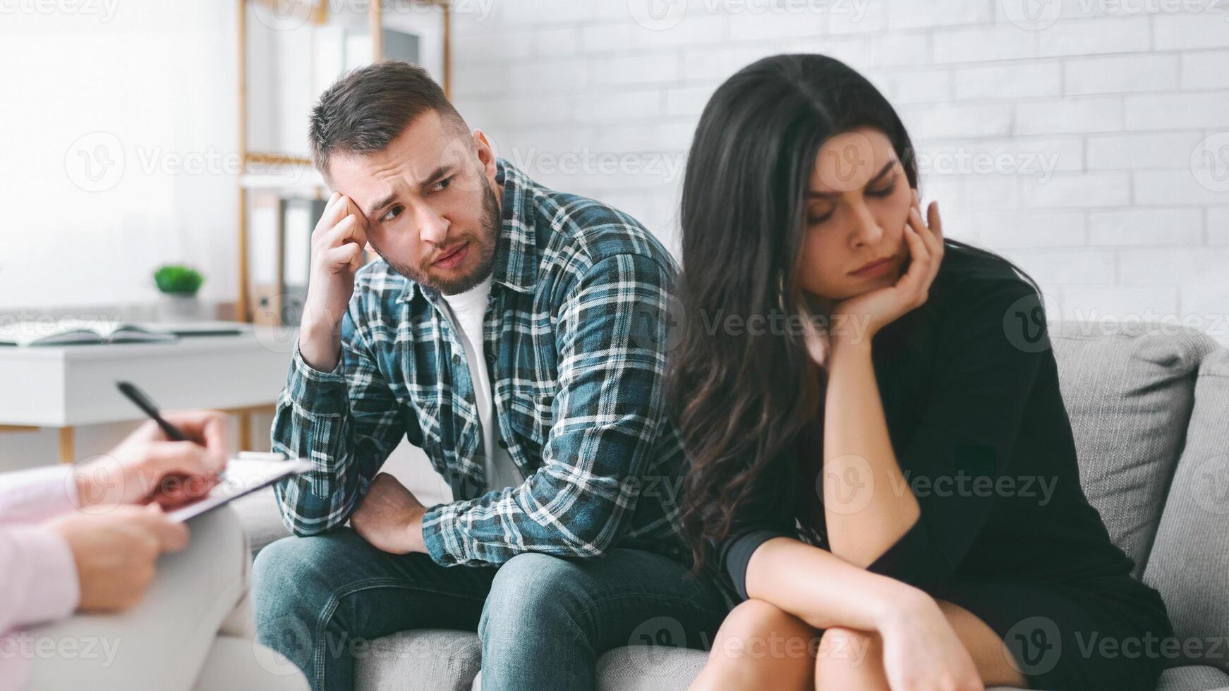A couple sits on a grey couch in a therapy session, looking down with expressions of sadness or worry. A therapists hand is visible in the foreground, holding a pen and notepad photo