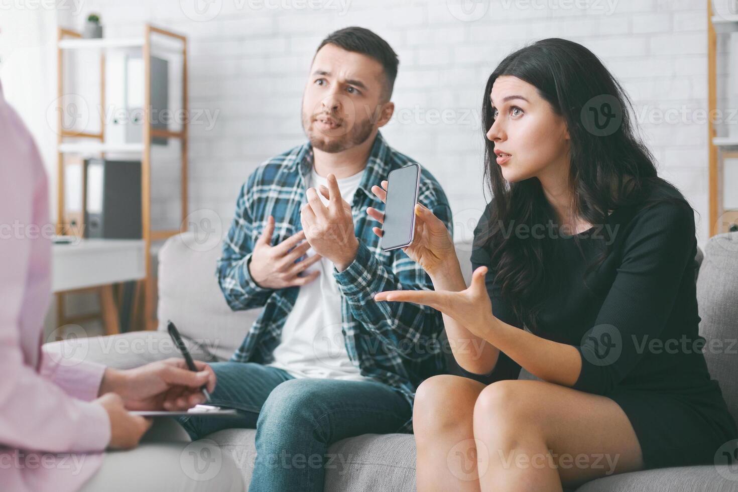 A young couple sits on a couch during a therapy session. They are discussing their smartphone usage, with the woman holding a smartphone in her hand and pointing to it with her left hand photo