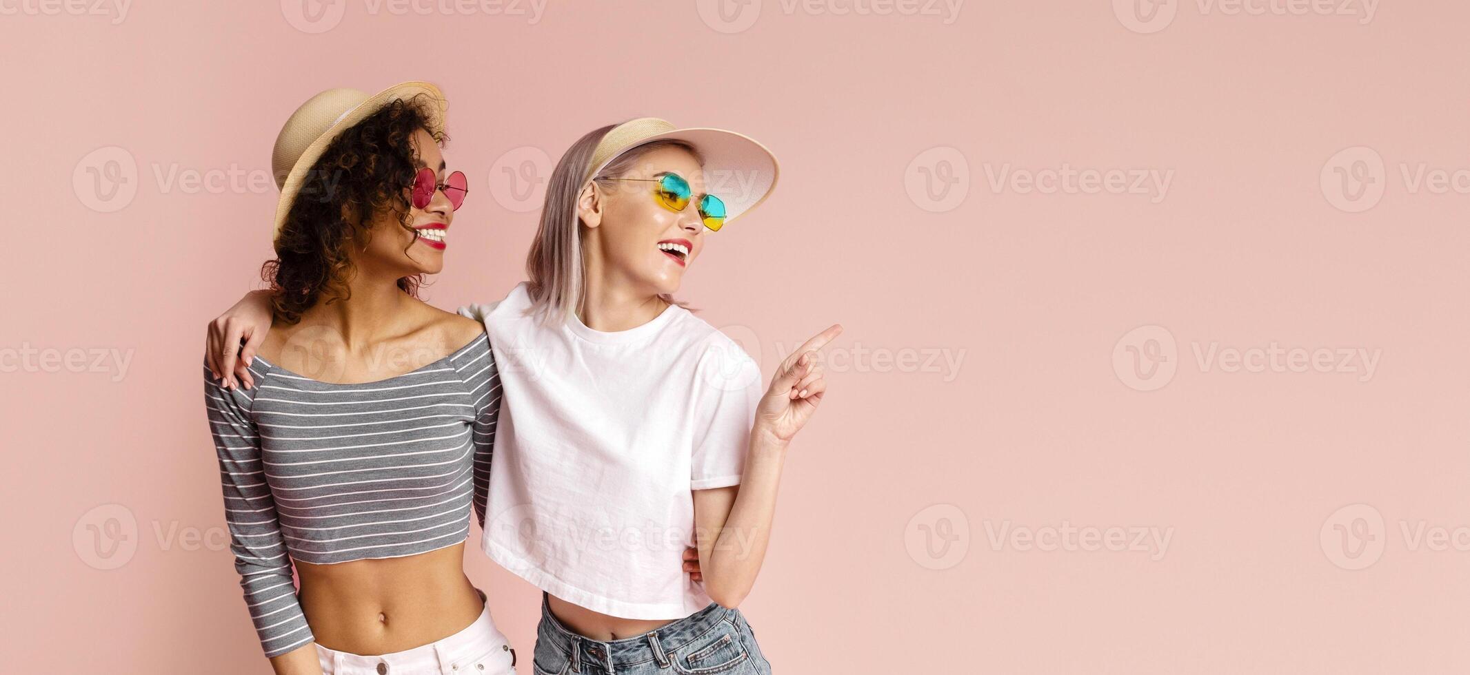 Two young women are standing in front of a solid pink backdrop. Both women are wearing sunglasses and smiling. The woman on the right is pointing to the left of the frame, copy space photo
