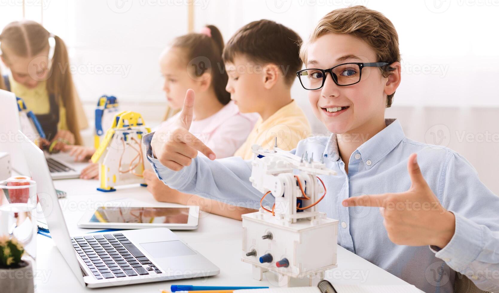 A young boy sits in a classroom setting with other students. He is smiling and pointing at a small robot he has built, demonstrating his learning and enthusiasm for STEM education. photo