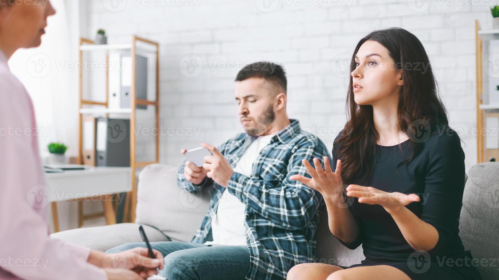 Spouses sit on a couch in an office, receiving counseling from a therapist. The woman is speaking, gesturing with her hands, while her partner appears distracted, looking at his phone. photo