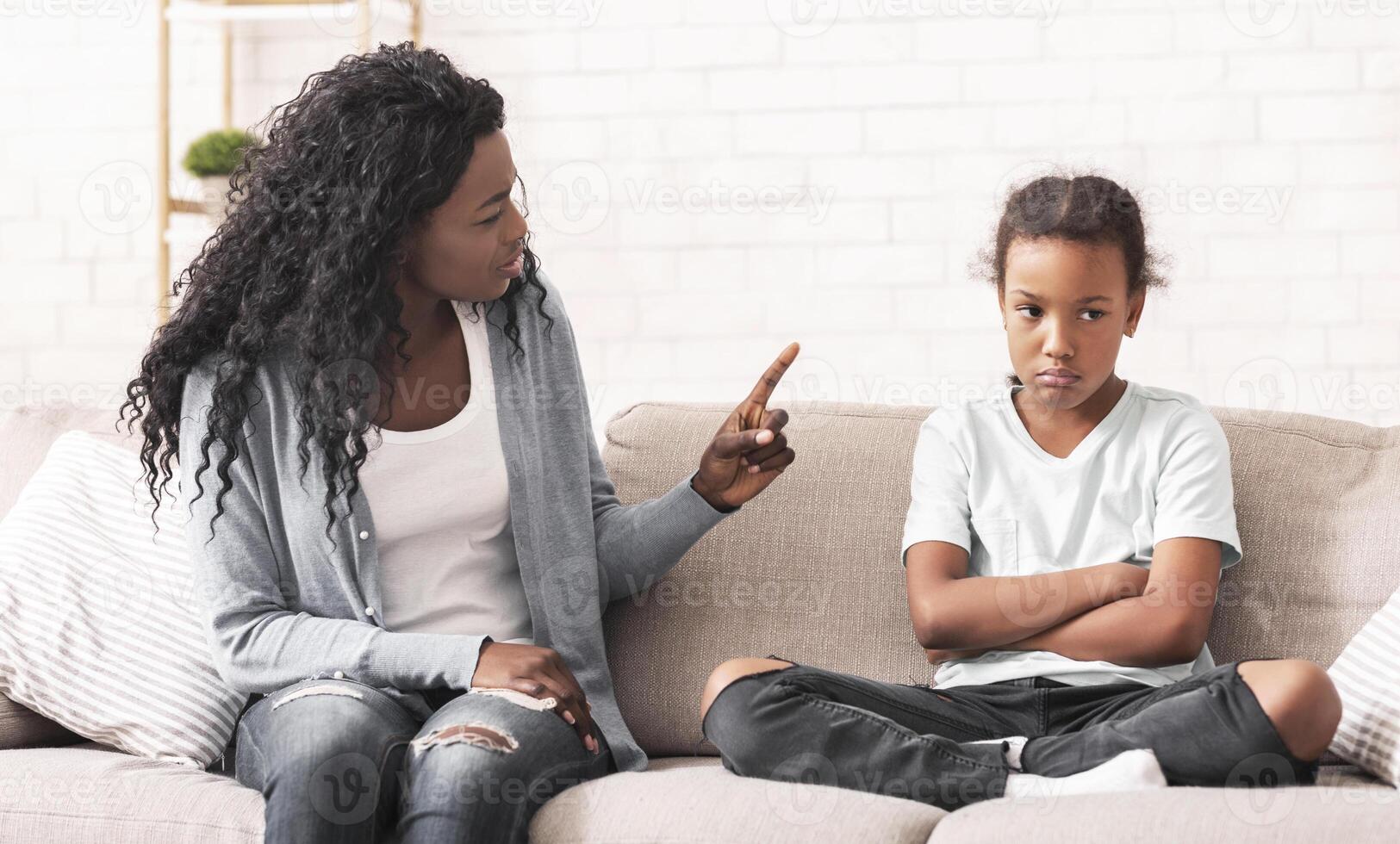 Family Discipline Concept. Grumpy black mom scolding her naughty little daughter on sofa at home photo