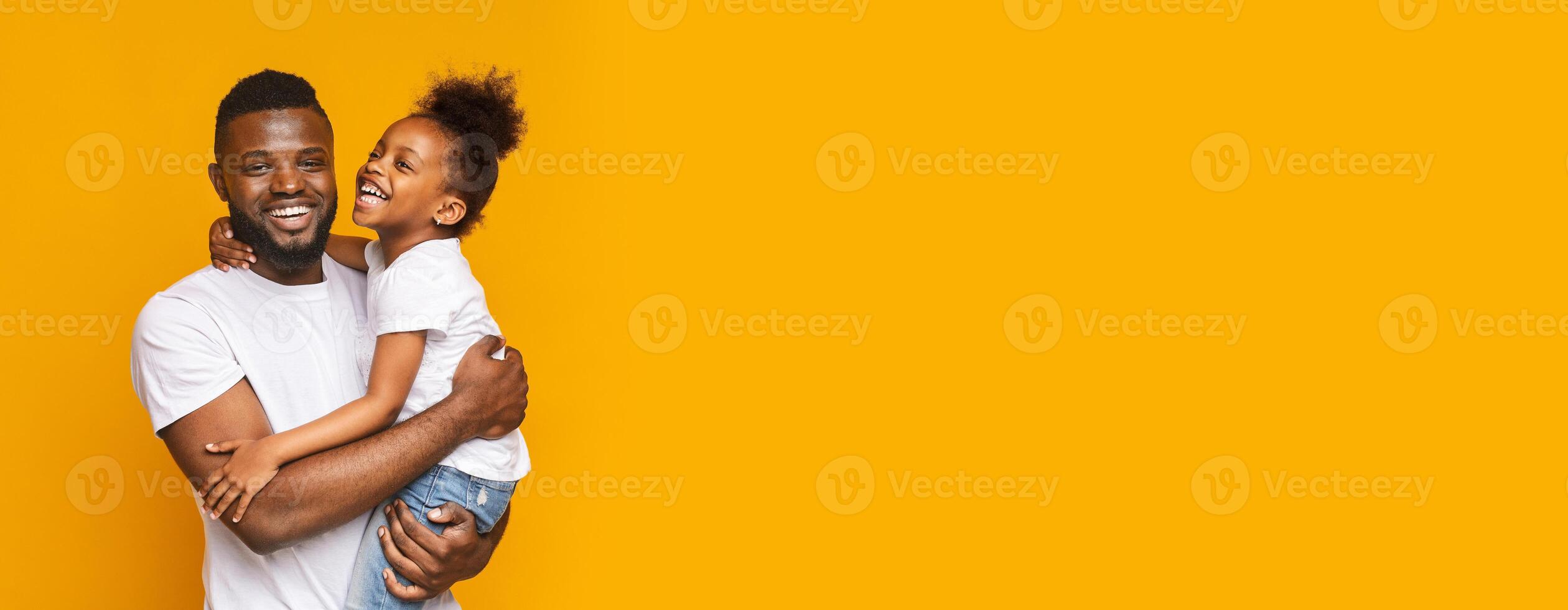 A cheerful black father holds his smiling daughter in his arms, both radiating happiness. The backdrop features a vibrant yellow color, enhancing their joyful expressions, copy space photo