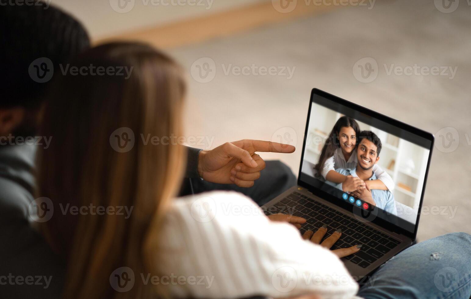 A couple is sitting on a couch at home, chatting with their friends on a laptop. The man is pointing at the screen, while the woman is smiling and looking at the laptop photo