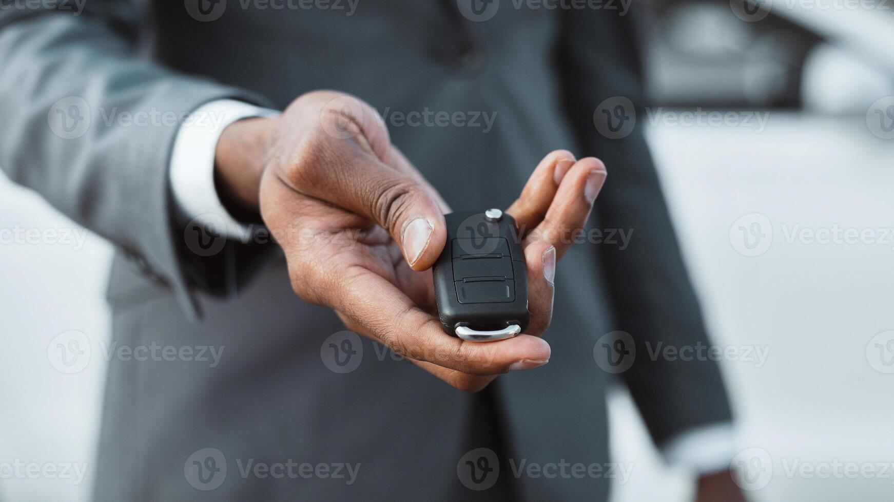 Unrecognizable man in a gray suit holds out a car key, showing the silver key fob with multiple buttons. The man is standing in front of a white car, with the cars exterior out of focus. photo