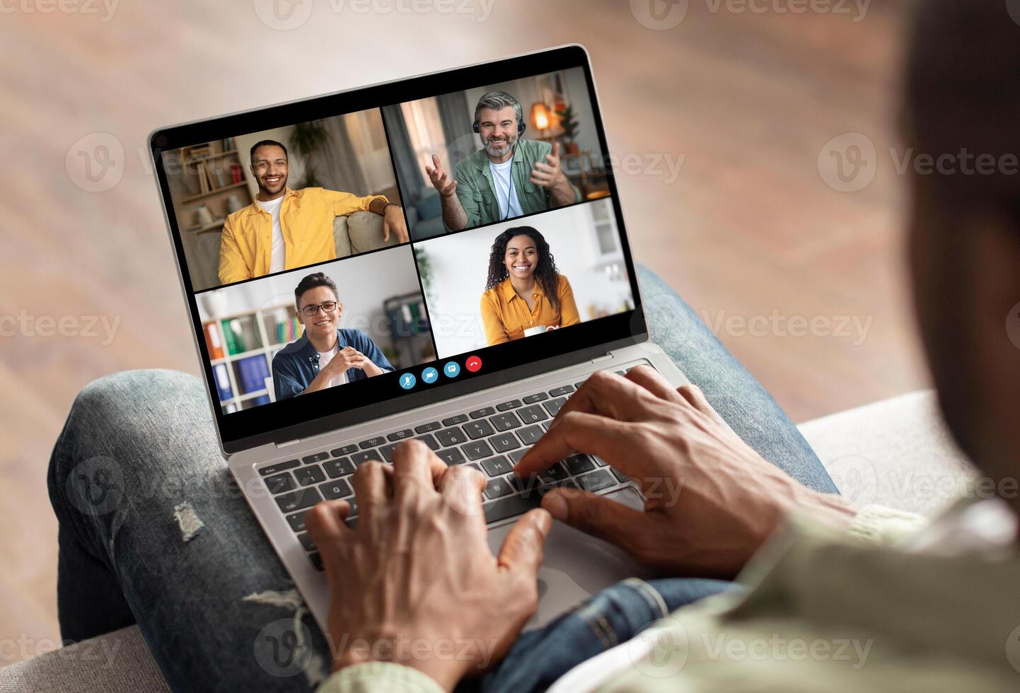 African American man sitting on a couch, using his laptop to participate in an online conference. There are four people visible on the laptop screen, and the man is typing on the keyboard. photo