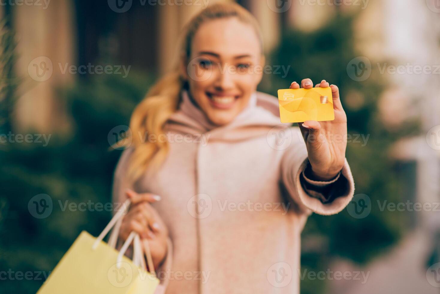 A young woman wearing a pink coat stands outside, holding a credit card in her right hand and a shopping bag in her left. She is smiling at the camera. She appears to be happy photo