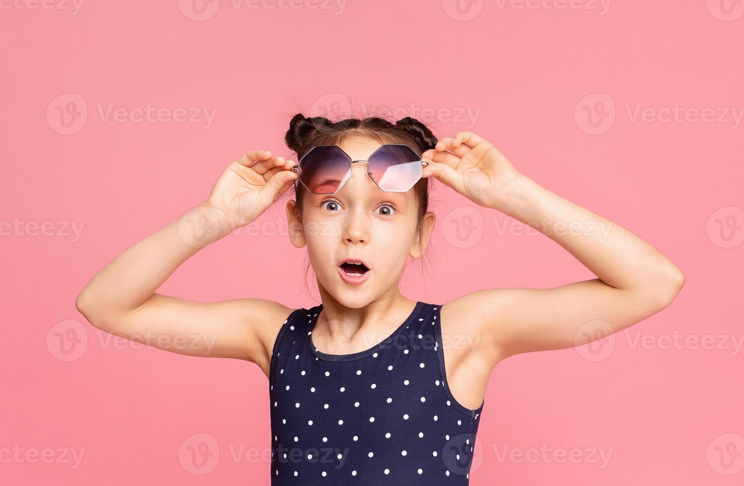 A young girl showcases her excitement while wearing a polka dot swimsuit and holding sunglasses. She stands against a vibrant pink background, appearing animated and playful. photo