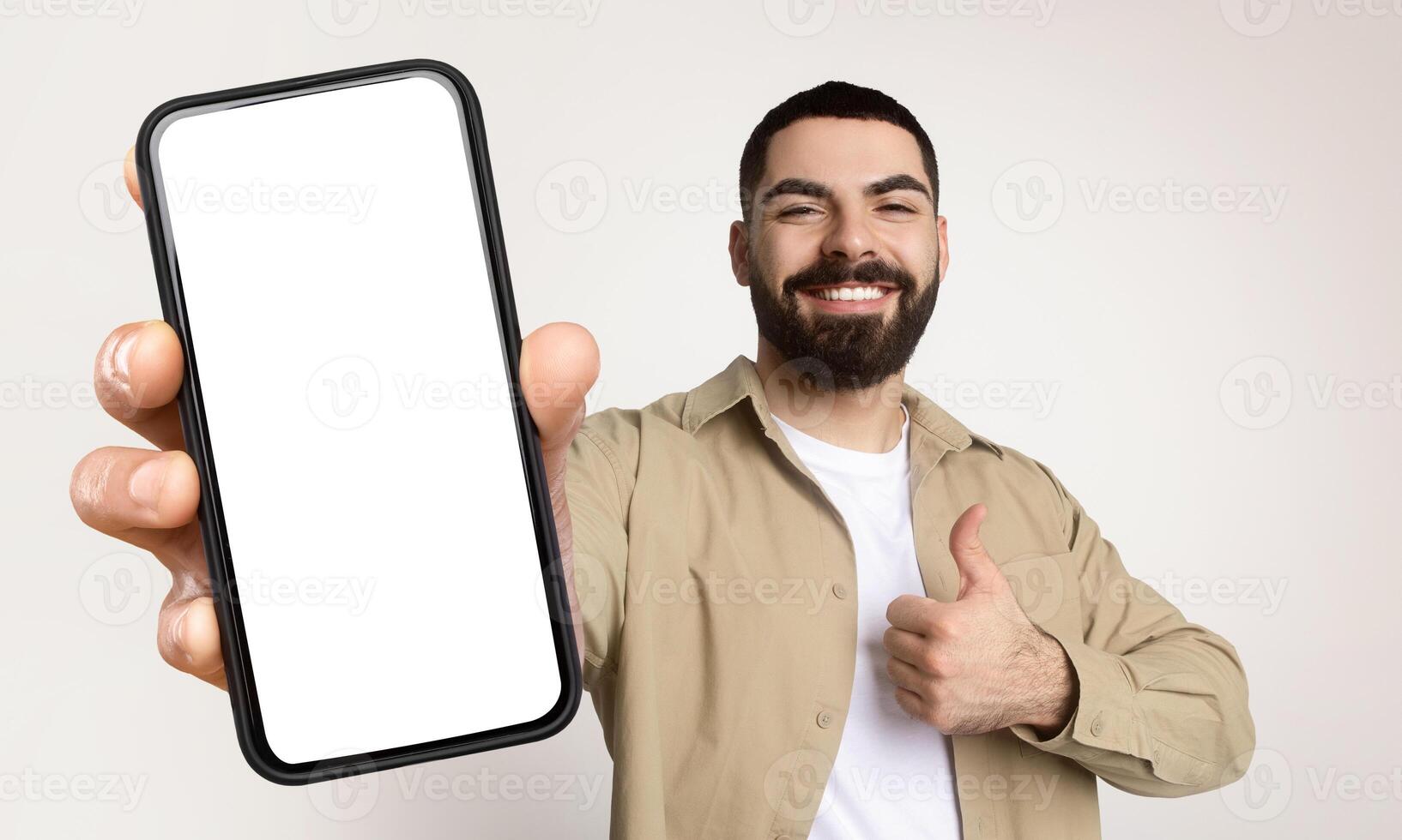 Millennial man is holding a smart phone with white blank screen in his hand, showing thumb up, isolated on beige studio background photo