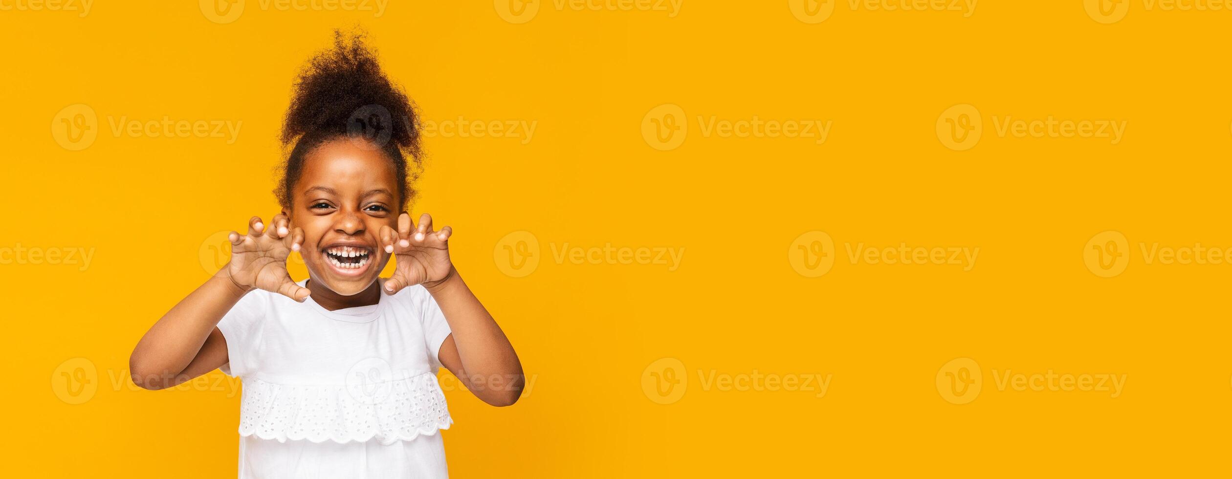 A young black girl with curly hair expresses joy and playfulness by making funny faces while standing against a bright yellow backdrop. Her laughter and cheerful demeanor create a lively atmosphere photo