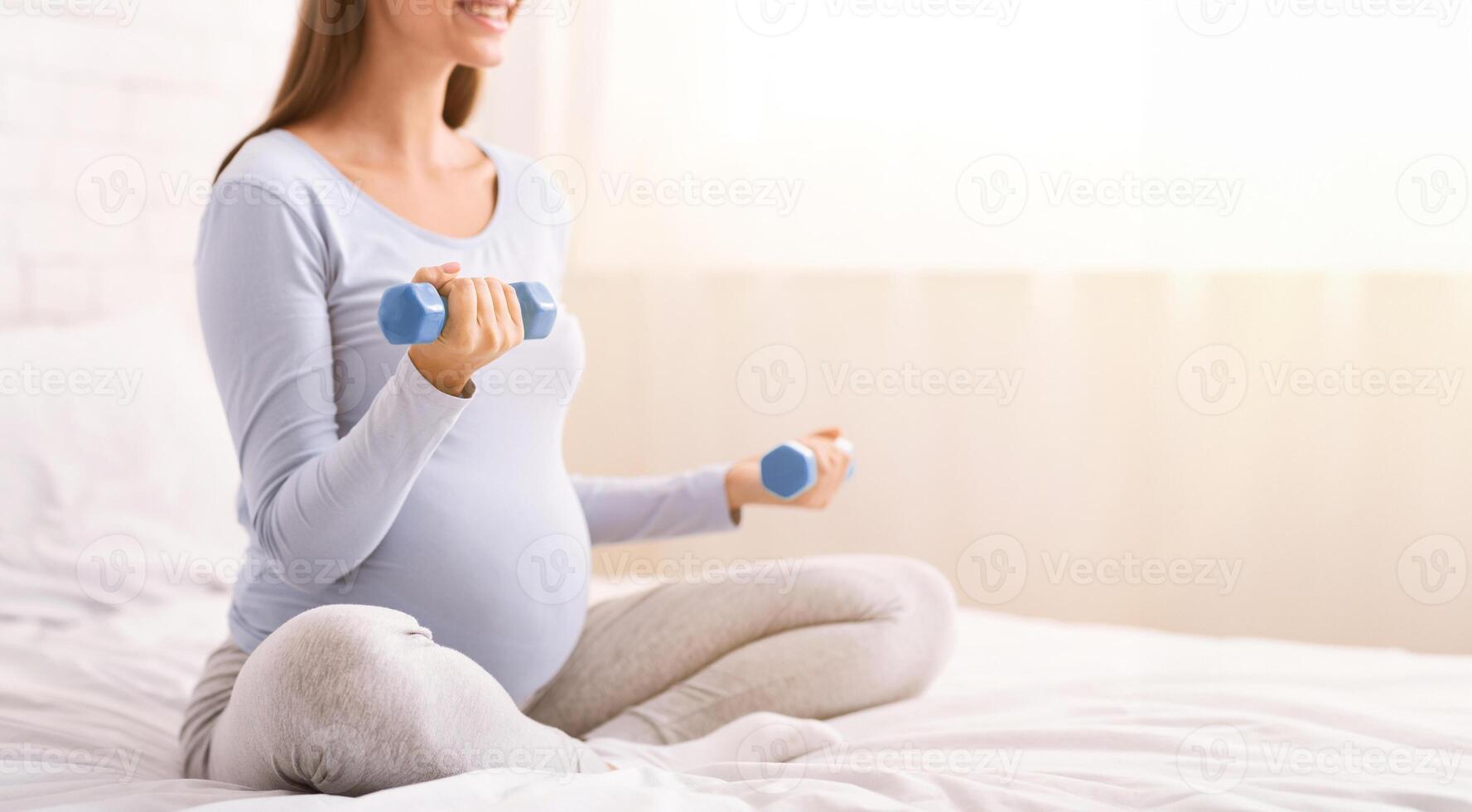 Unrecognizable Pregnant Girl Exercising With Dumbbells Sitting On Bed At Home. Workout And Pregnancy. Cropped photo