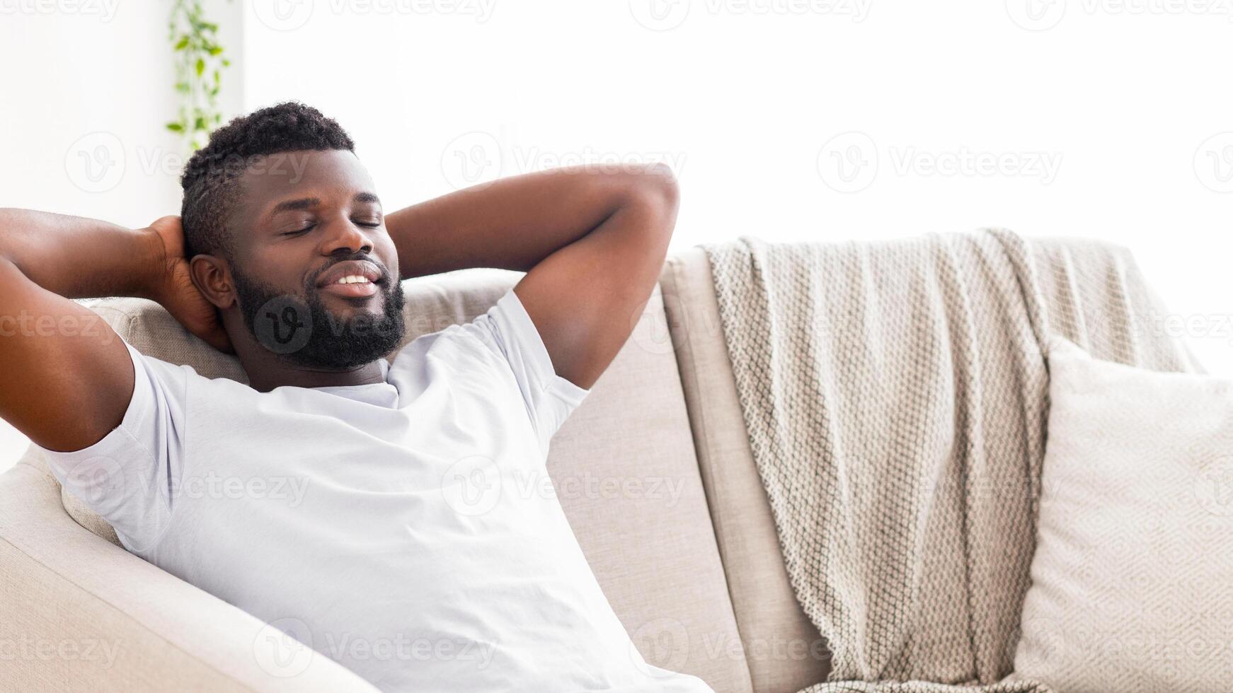 This image shows a black man with a beard relaxing on a white couch. He has his arms behind his head and his eyes are closed as he leans back into the cushions, copy space photo