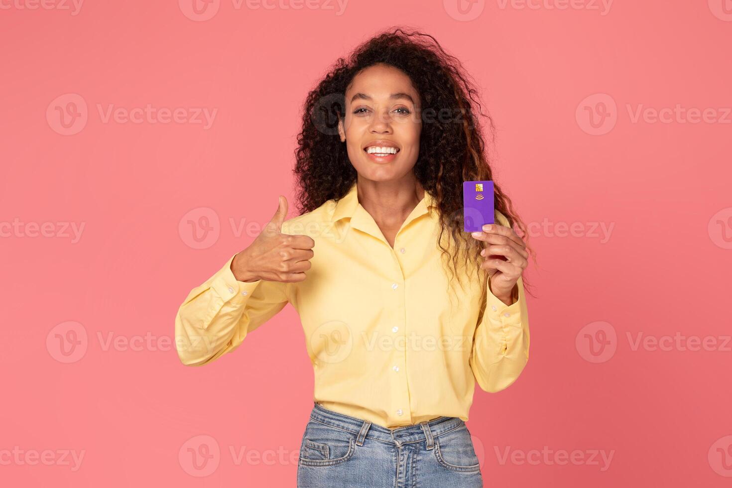 African American young woman with curly hair is smiling and giving a thumbs up while holding a purple credit card in her other hand. The background is a solid pink. photo