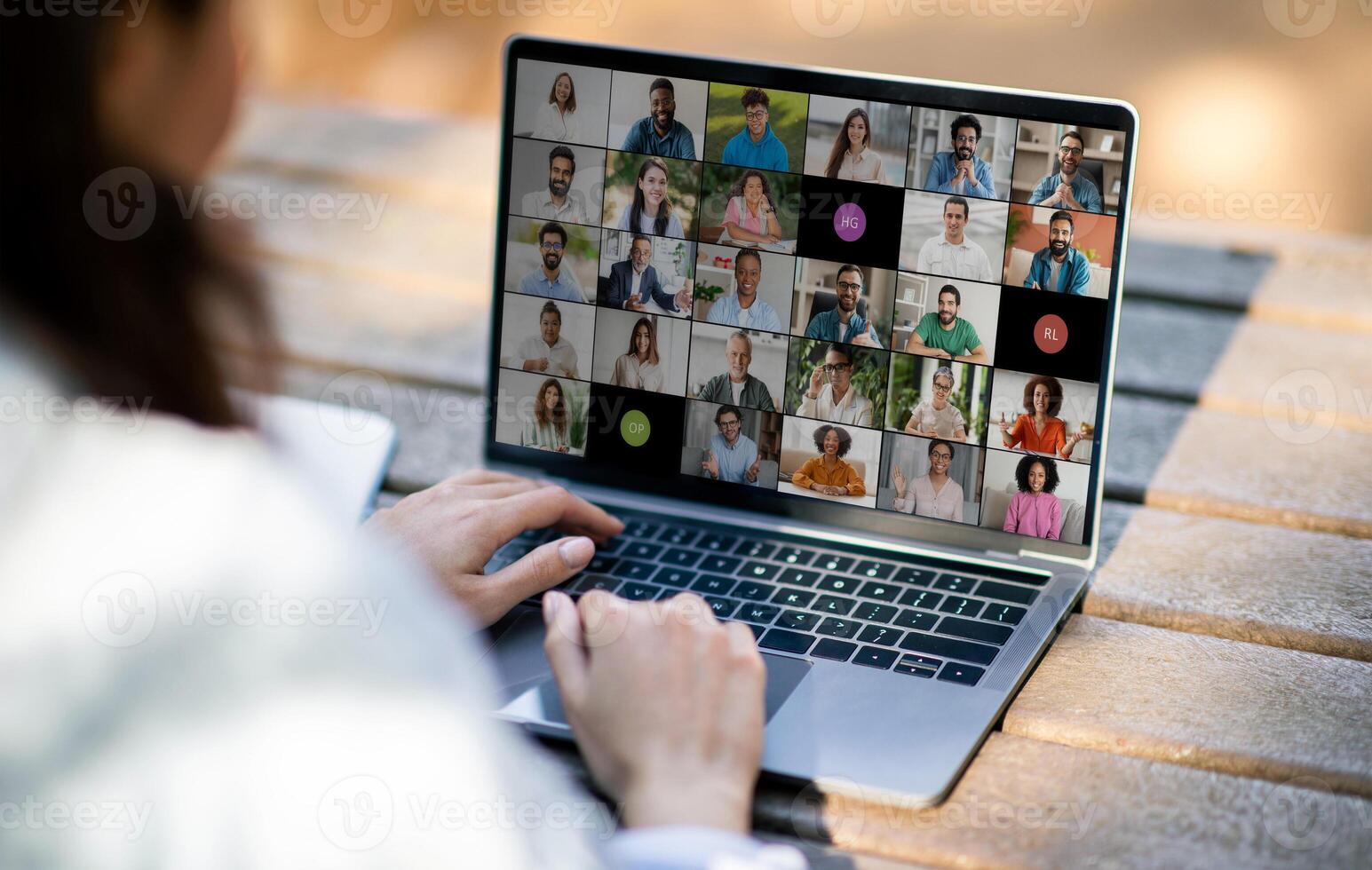 Lady sitting at an outdoor table, actively engaging in a virtual business meeting on a laptop. The screen displays numerous participants in different windows. photo