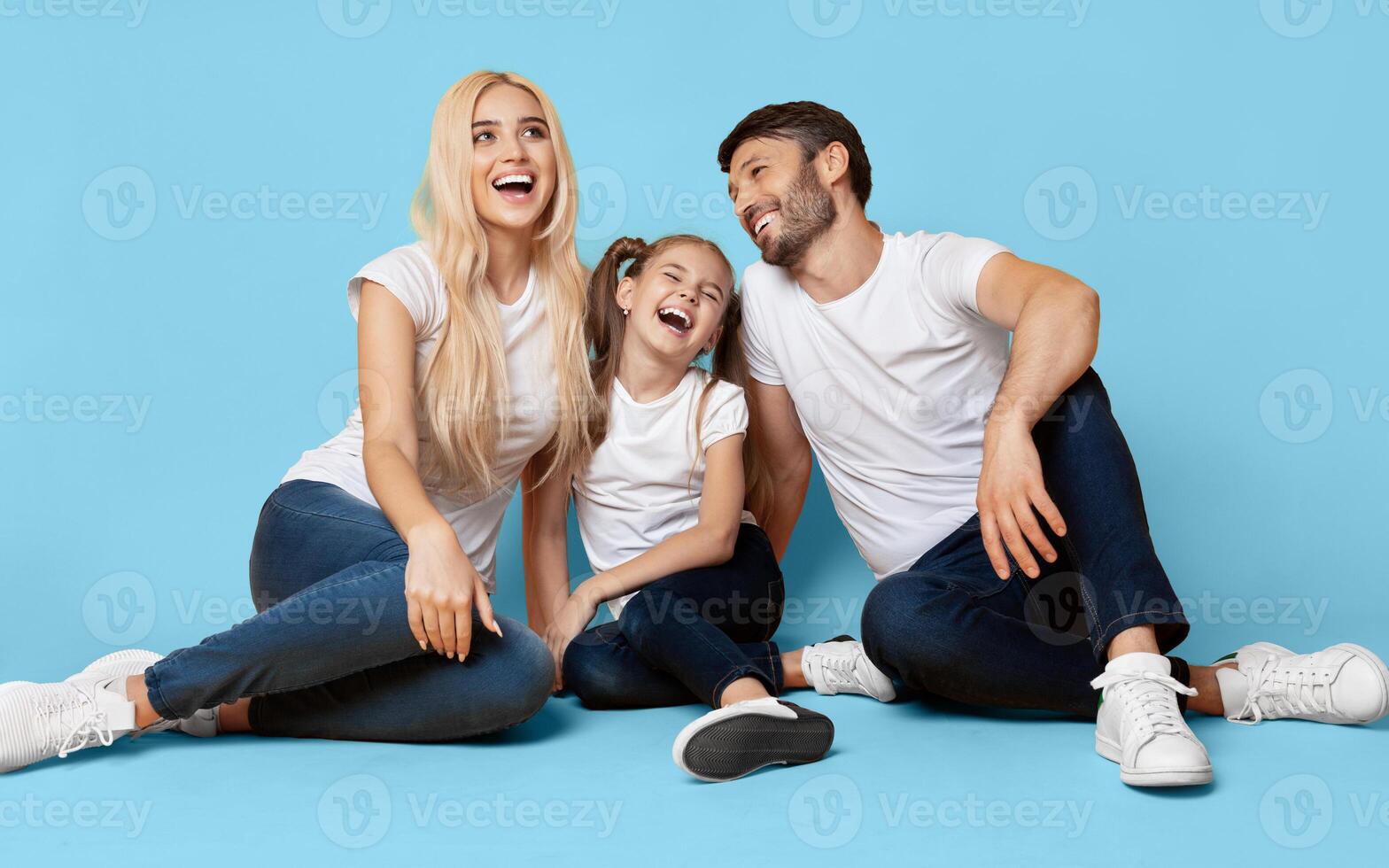 A family of three, a man, a woman, and a young girl, are sitting on a blue backdrop. The family is laughing and looking happy, with the girl in the middle smiling wide. photo