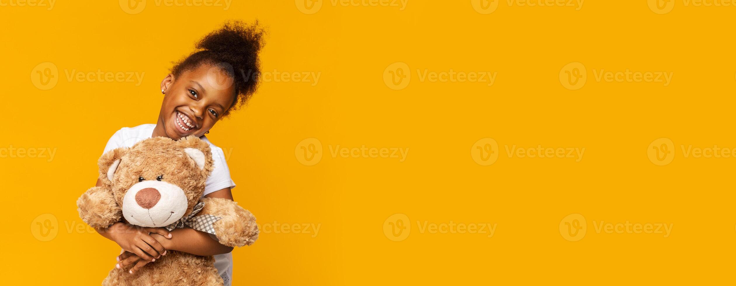 A joyful young black girl smiles widely while hugging a large teddy bear. She stands against a vibrant yellow backdrop, showcasing her excitement and affection for her favorite toy, copy space photo