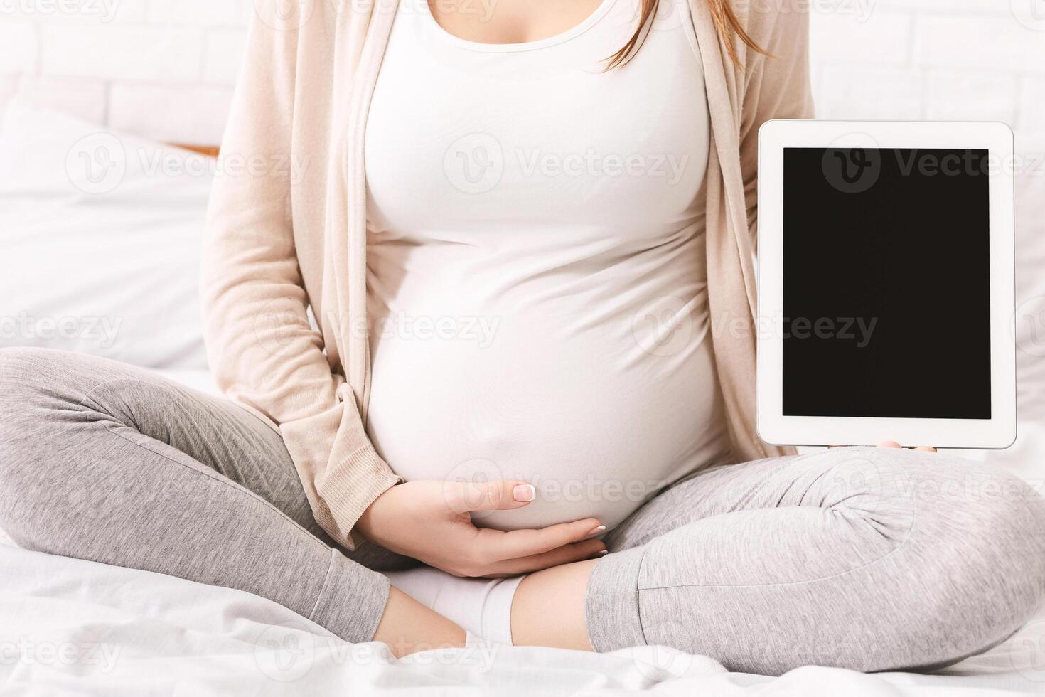 A pregnant woman is sitting on a bed, holding a tablet with black screen in her hands. She appears focused on the screen as she interacts with the device, cropped photo