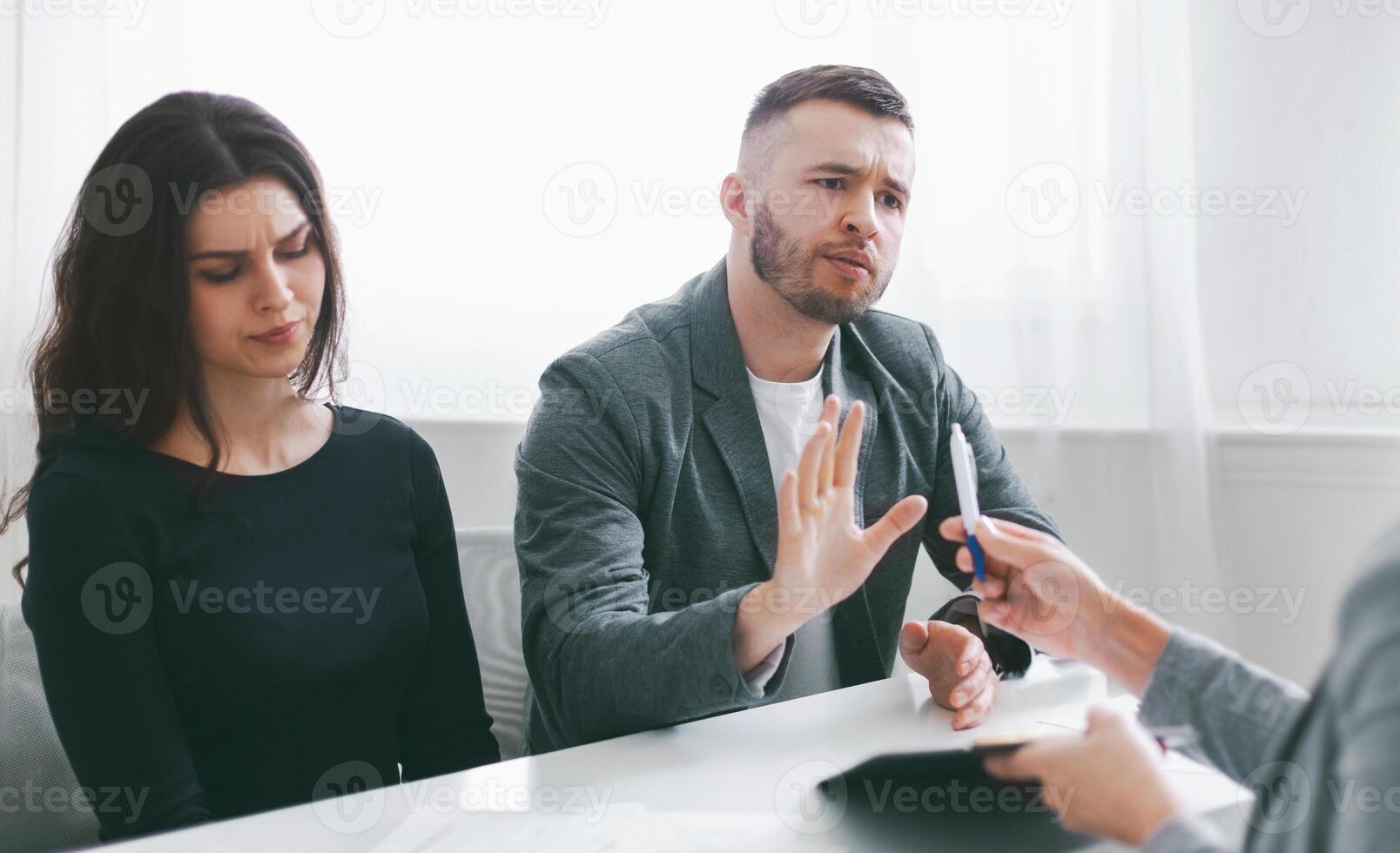 A couple sits at a table in a meeting with a lawyer, with the man gesturing and refusing to sign papers being handed to them. The woman sits beside the man, with a concerned expression on her face. photo