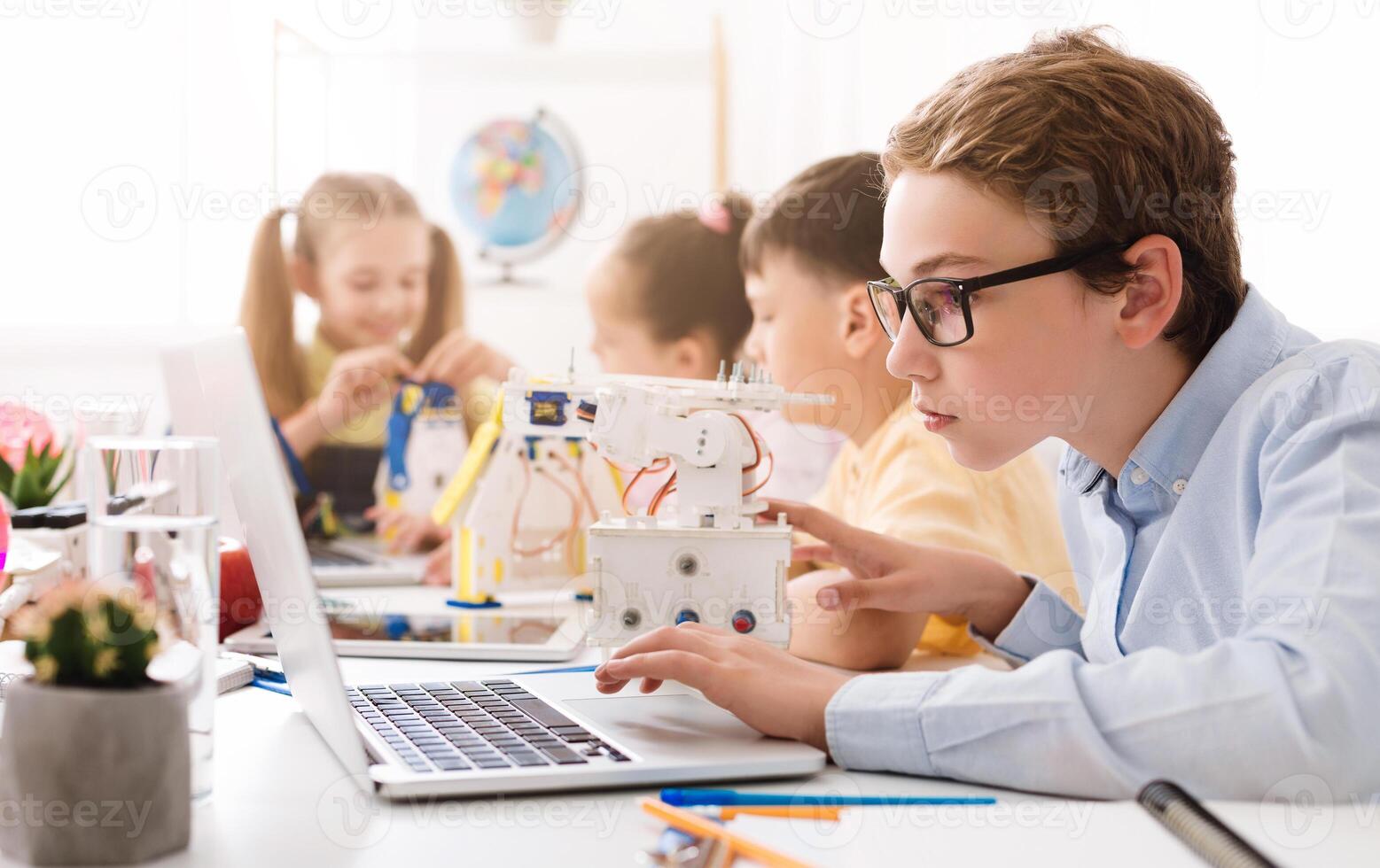 A teenage boy wearing glasses sits at a desk in a classroom, using a laptop computer. He is surrounded by other students, including a girl who appears to be working on a small robot. photo