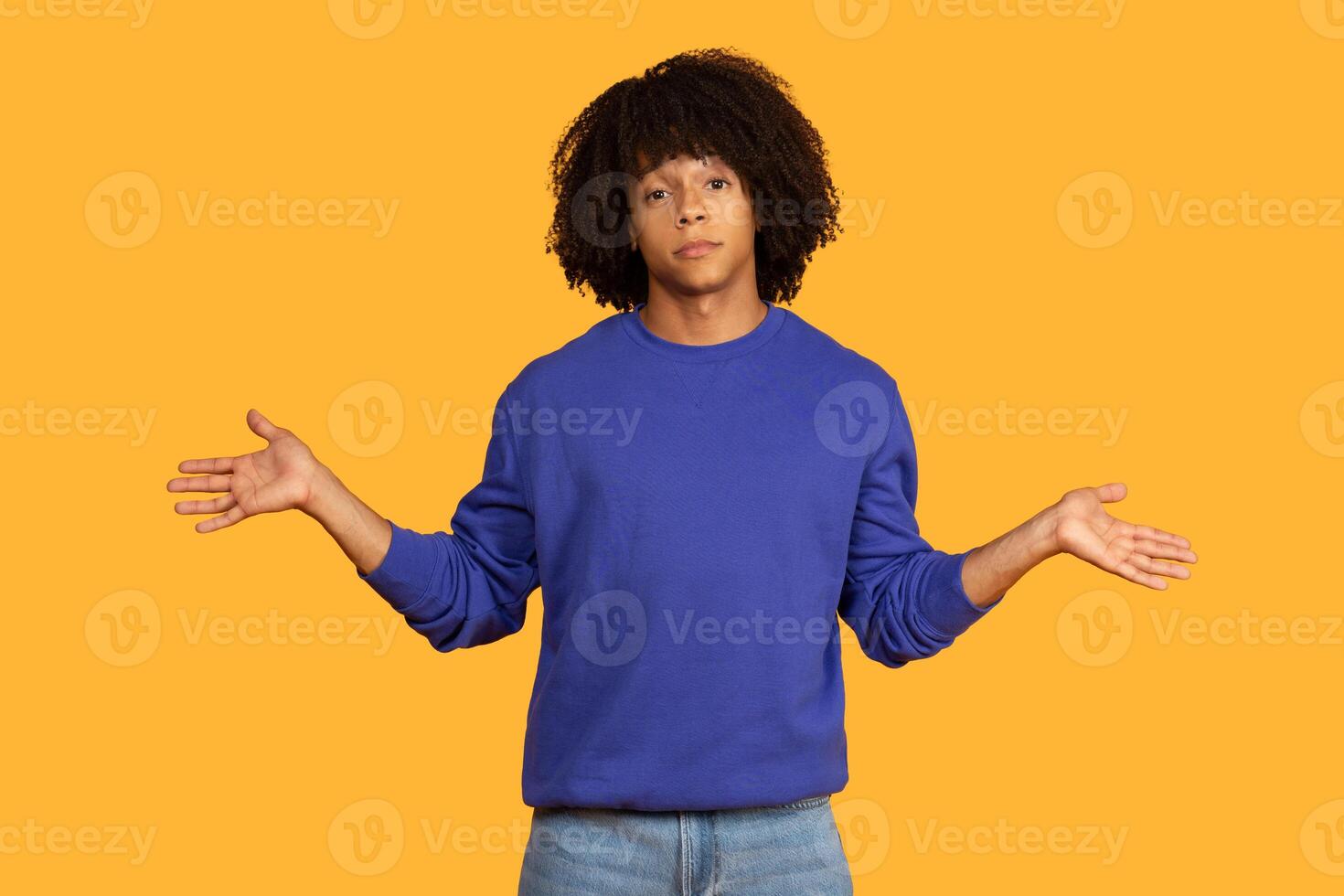 A young man with an curly hairstyle is standing in front of a yellow background. He is wearing a blue sweater and jeans. He is gesturing with both hands, palms facing outwards photo