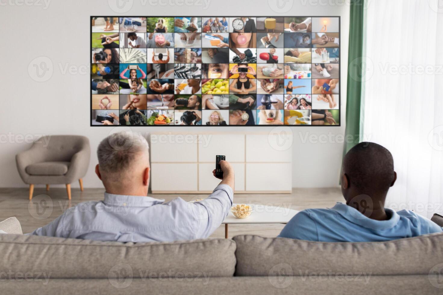 Two men are seated on a couch in a living room, watching television. The television is displaying a grid of images, and the man on the left is holding a remote control. photo