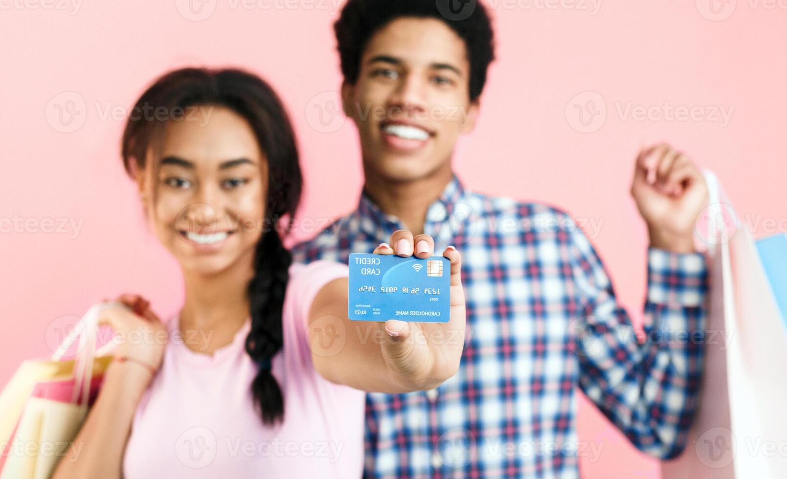 Consumerism concept. Teenage couple showing credit card and holding shopping bags, pink background photo