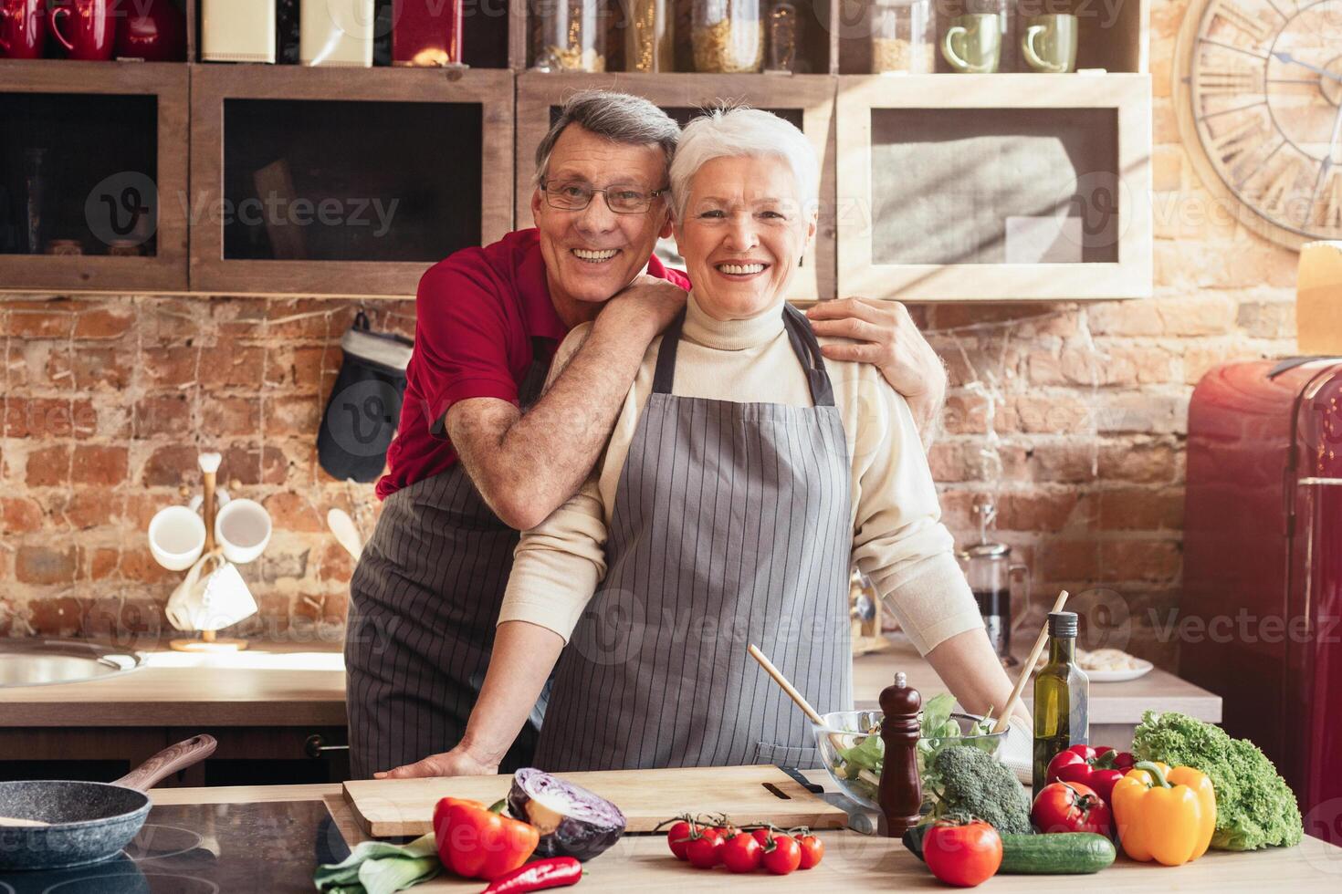 A senior couple smiles while preparing a meal in their kitchen. The woman is leaning forward, her hand resting on the countertop. There are fresh vegetables and a bowl of salad on the counter photo