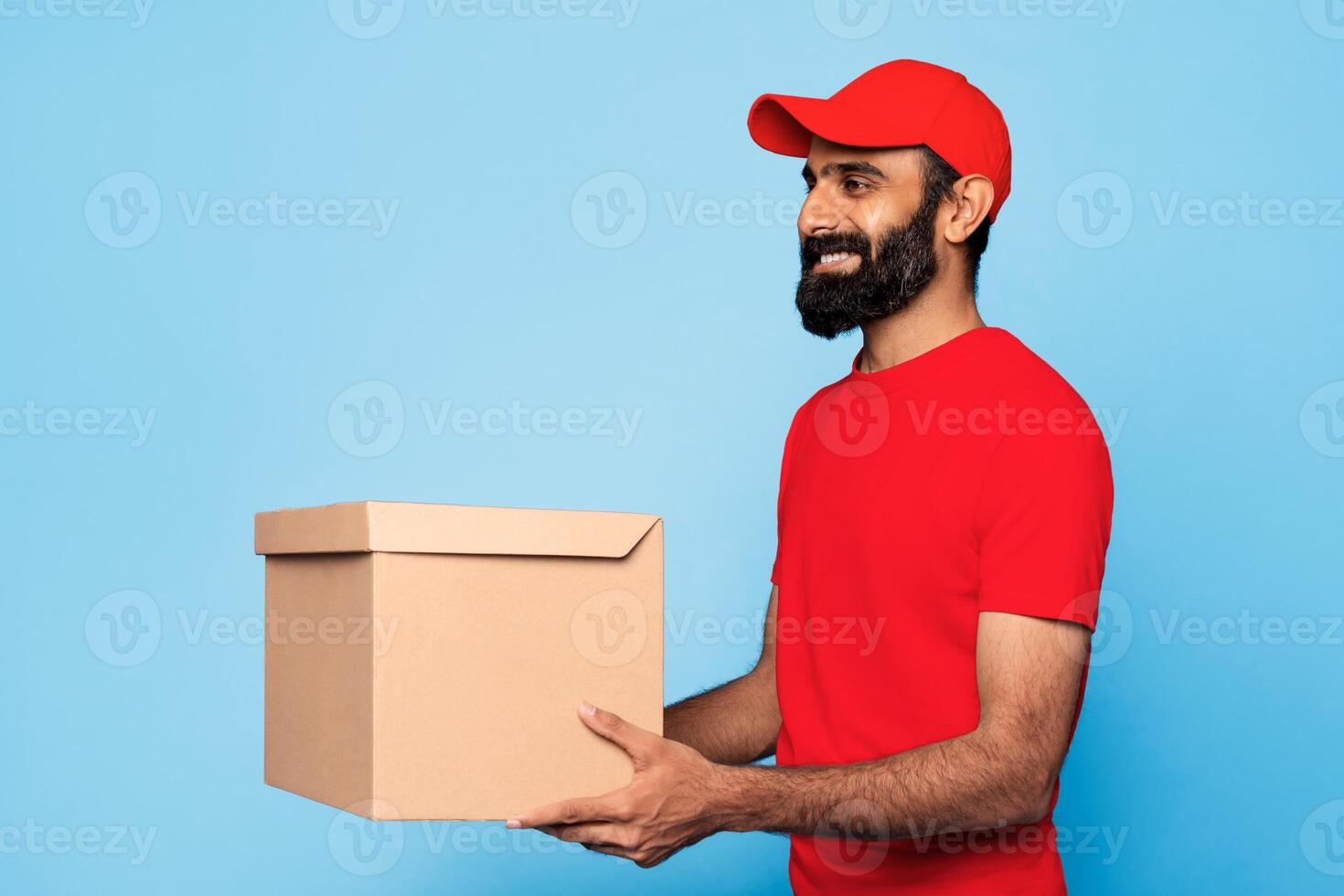 Indian man wearing a red t-shirt and a red cap is smiling and holding a brown cardboard box in his right hand. The man is standing in front of a blue background. photo