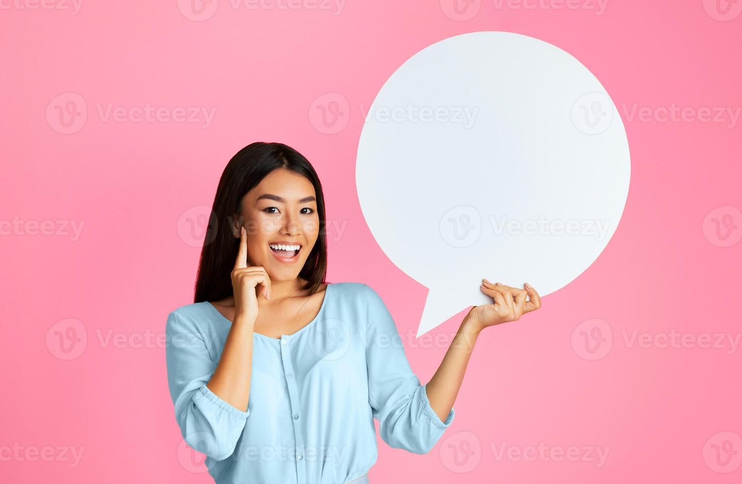 Asian smiling young woman standing in front of a pink background, holding a blank speech bubble in her right hand. Her left hand is touching her cheek photo