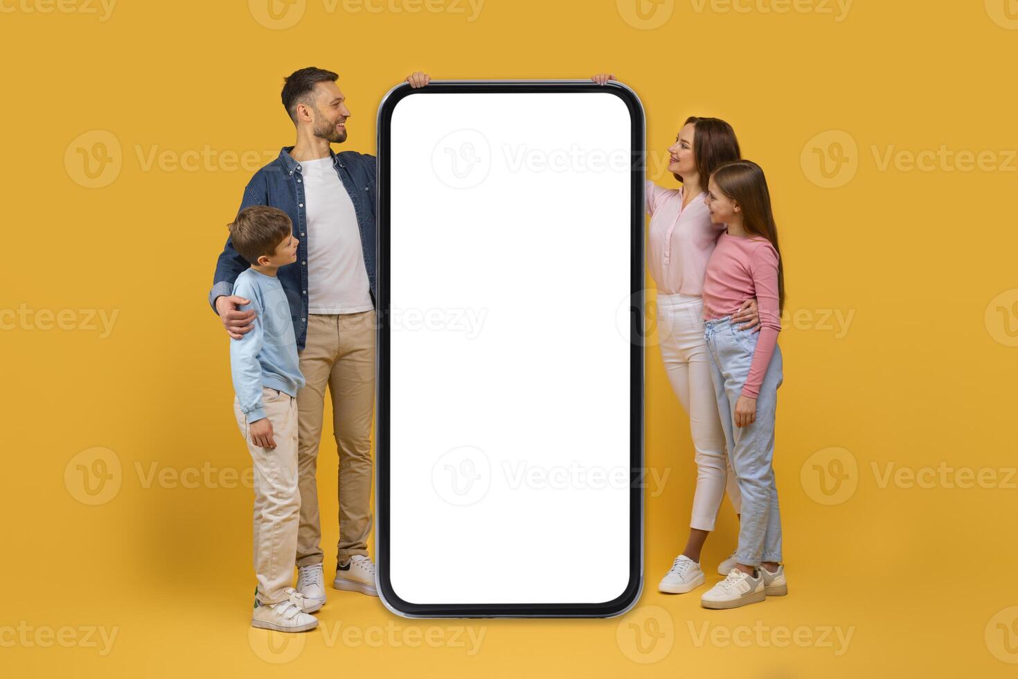 A family consisting of a man, woman, boy, and girl are standing together beside an oversized smartphone with a blank white screen, suitable for mockup purposes. photo