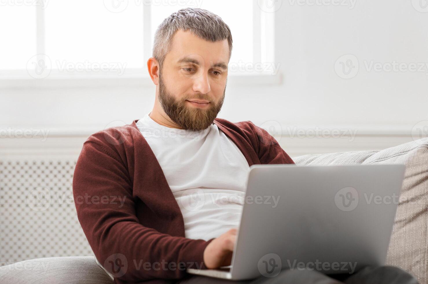 A man is seated on a couch, focusing intently on his laptop screen. His hands are busy typing on the keyboard as he works or browses online. photo