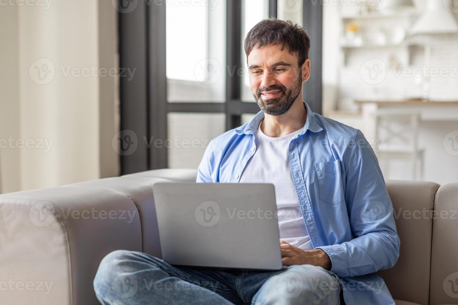 A man is sitting on a couch in a living room. He is wearing a blue shirt and jeans. He is looking at the laptop in his lap. He is smiling as he works. photo