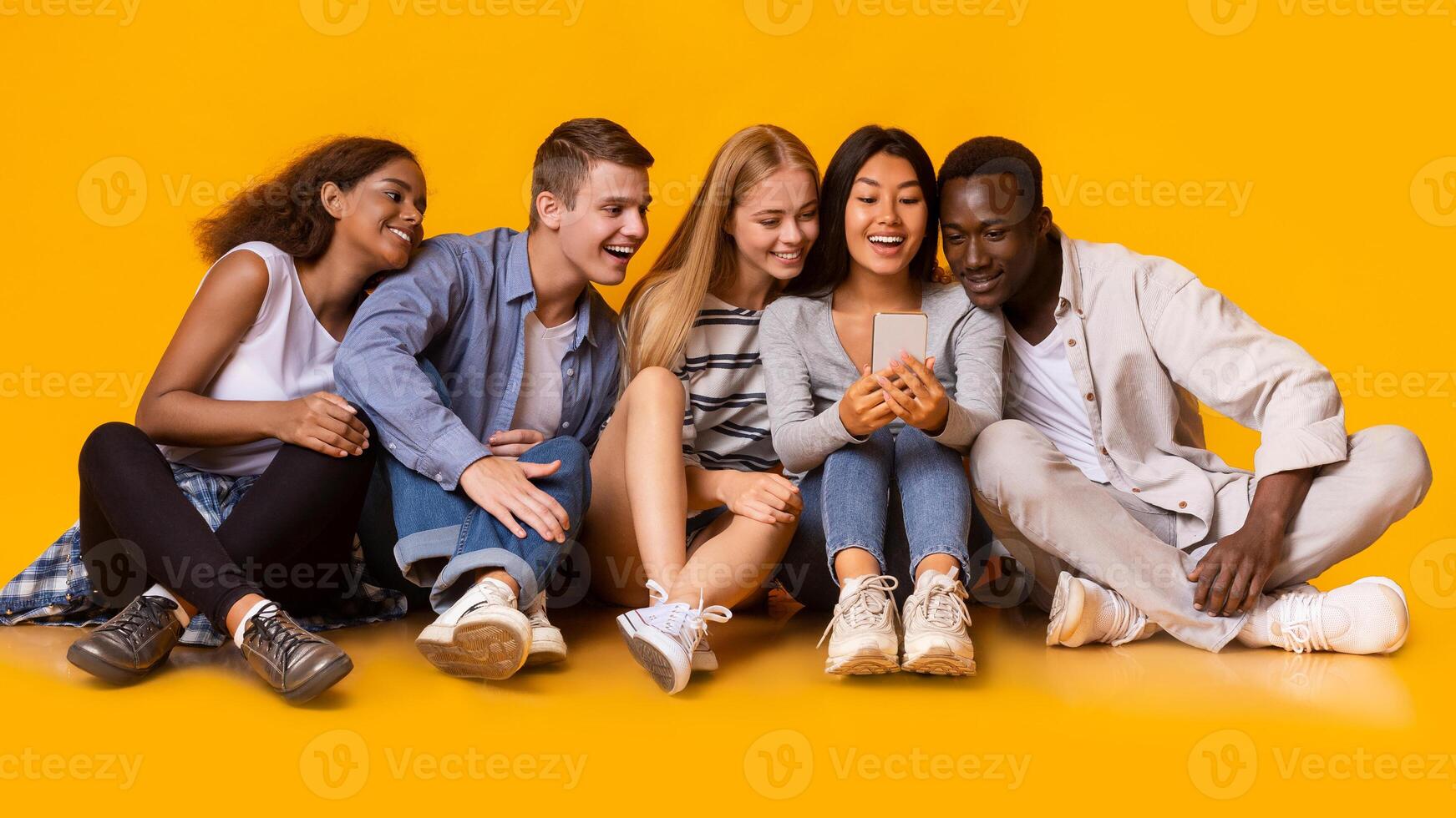 Group of multiracial cheerful classmates having break, watching videos on smartphone over yellow background, panorama photo