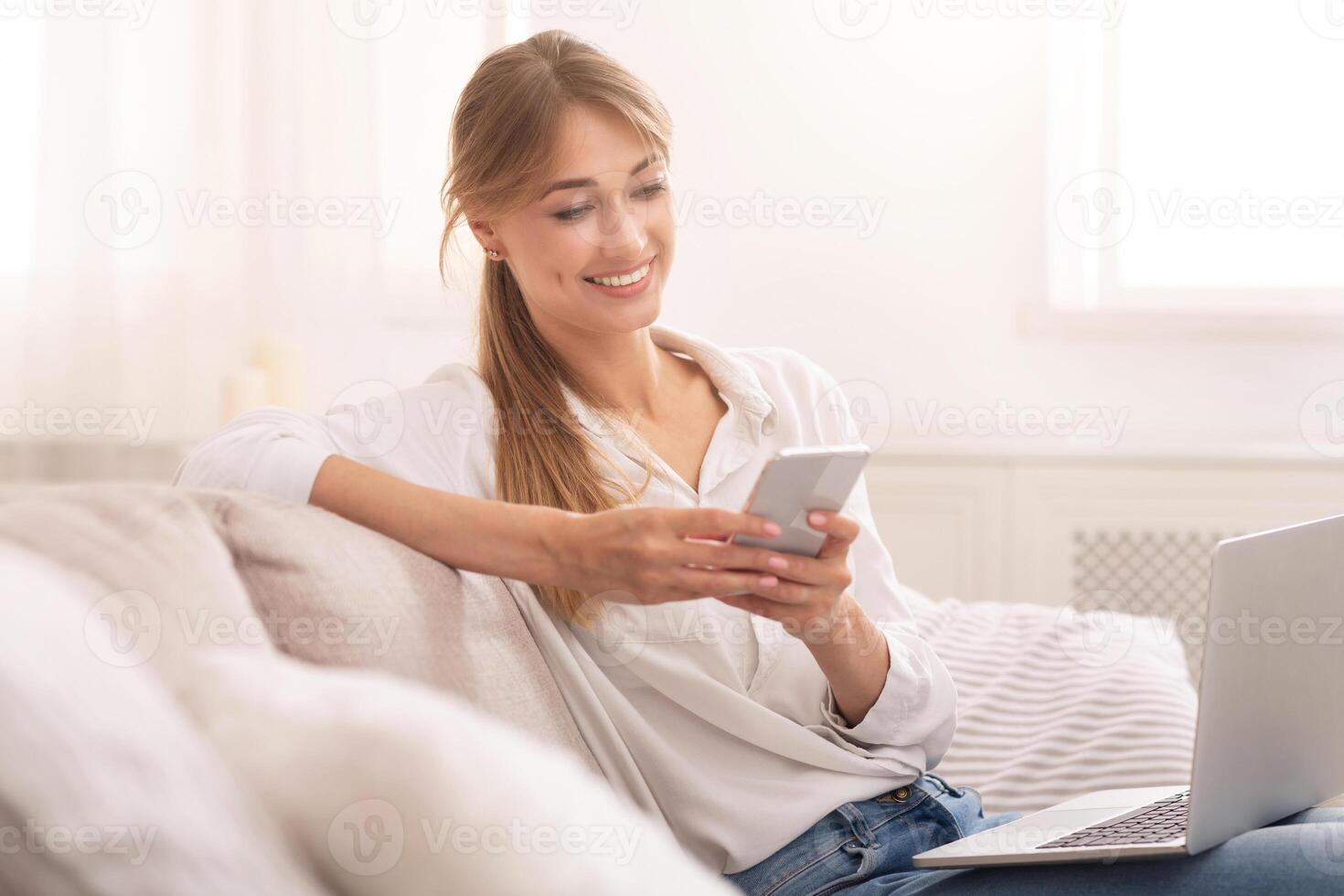Smiling Woman Using Mobile Phone And Laptop Relaxing Sitting On Couch At Home. Selective Focus photo