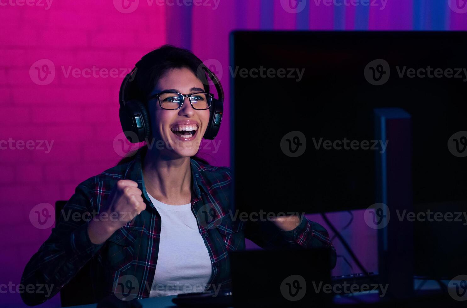 A young woman wearing glasses and headphones sits in front of a computer with a bright smile on her face. She is celebrating a victory while gaming in a room with vibrant purple and blue lighting photo