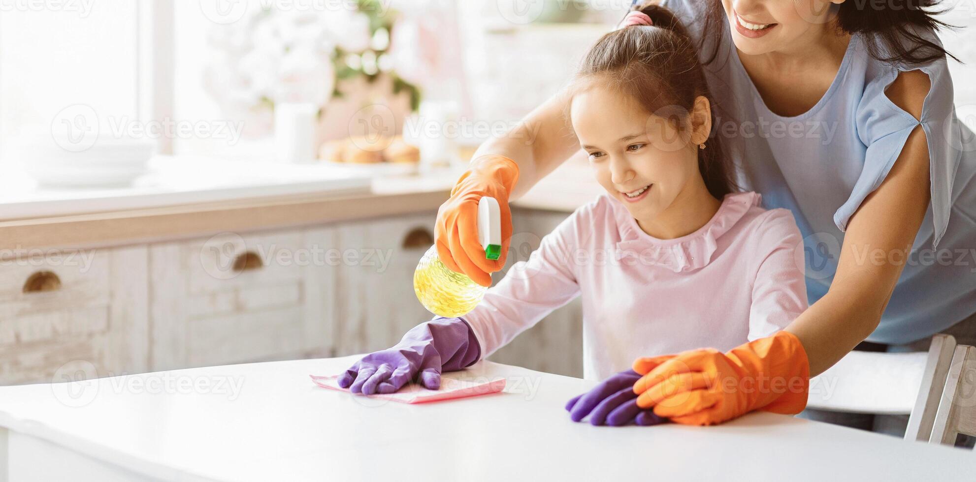 A young girl wearing pink shirt and purple gloves is cleaning a table in the kitchen while her mother, wearing a blue shirt and orange gloves, assists her, panorama with copy space photo
