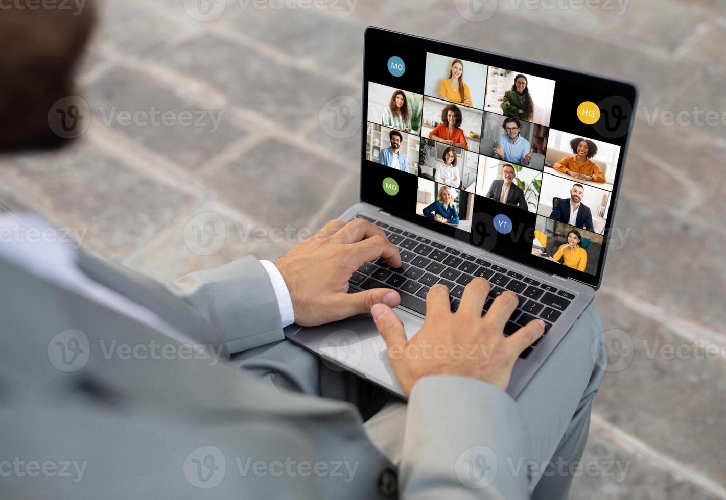 Black business professional in a grey suit is engaged in an online conference on a laptop while sitting outdoors on a stone-paved surface photo