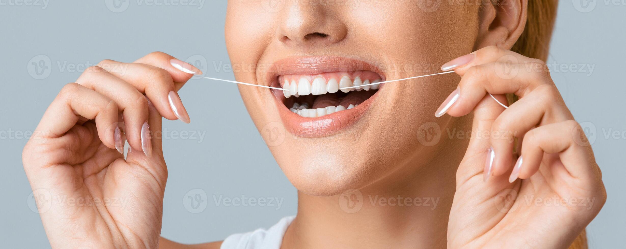 A close-up shot of a woman smiling while flossing her teeth with white floss against a grey background. Her teeth are white and bright, and she is clearly enjoying the process. photo