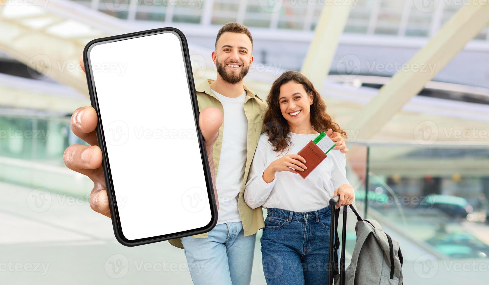 A beaming young man and woman stand inside a bustling airport terminal, showcasing a large, blank smartphone screen. photo