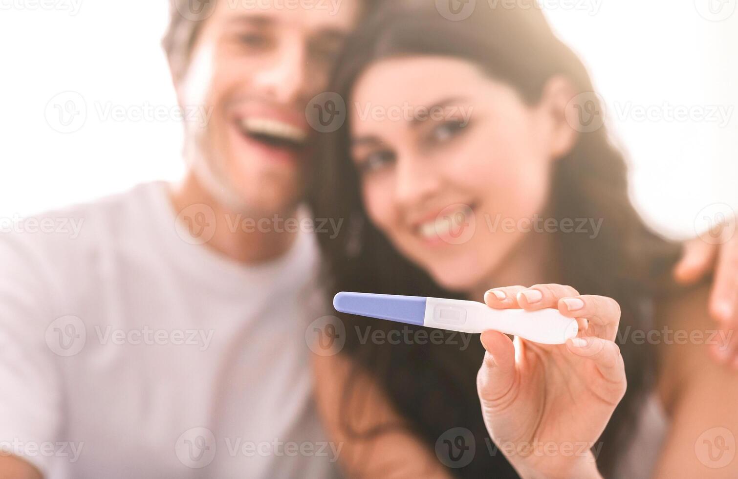 A couple is pictured holding a pregnancy test with a positive result. The woman is holding the test in front of the camera with a beaming smile, while the man is blurred in the background photo