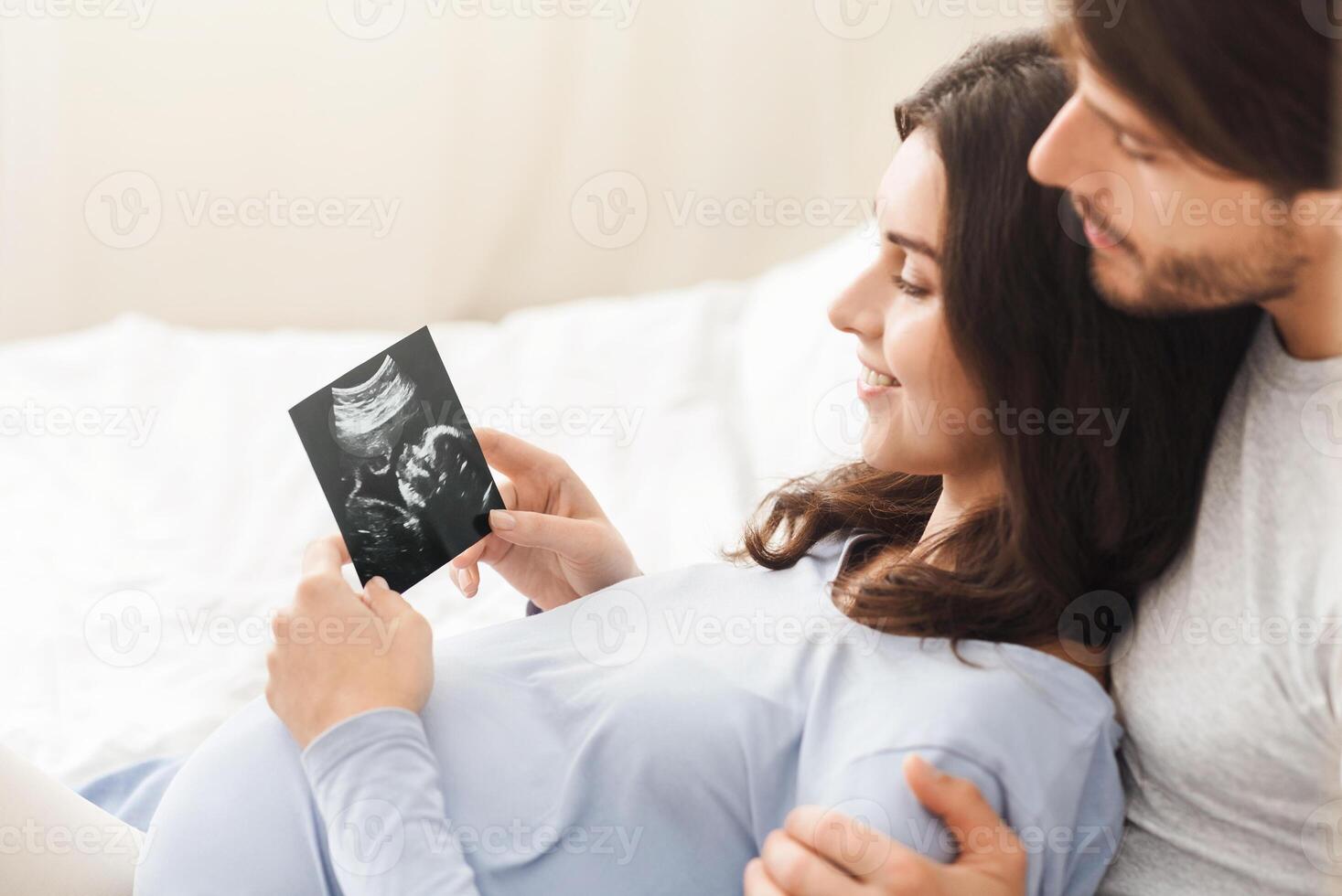 First portrait of our baby. Happy pregnant woman and her husband looking at sonogram picture, lying on bed at home, free space photo