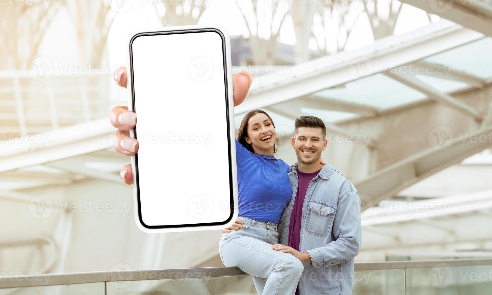 A man and woman are standing together, holding up a cell phone with white blank screen, mockup copy space. Traveling app photo