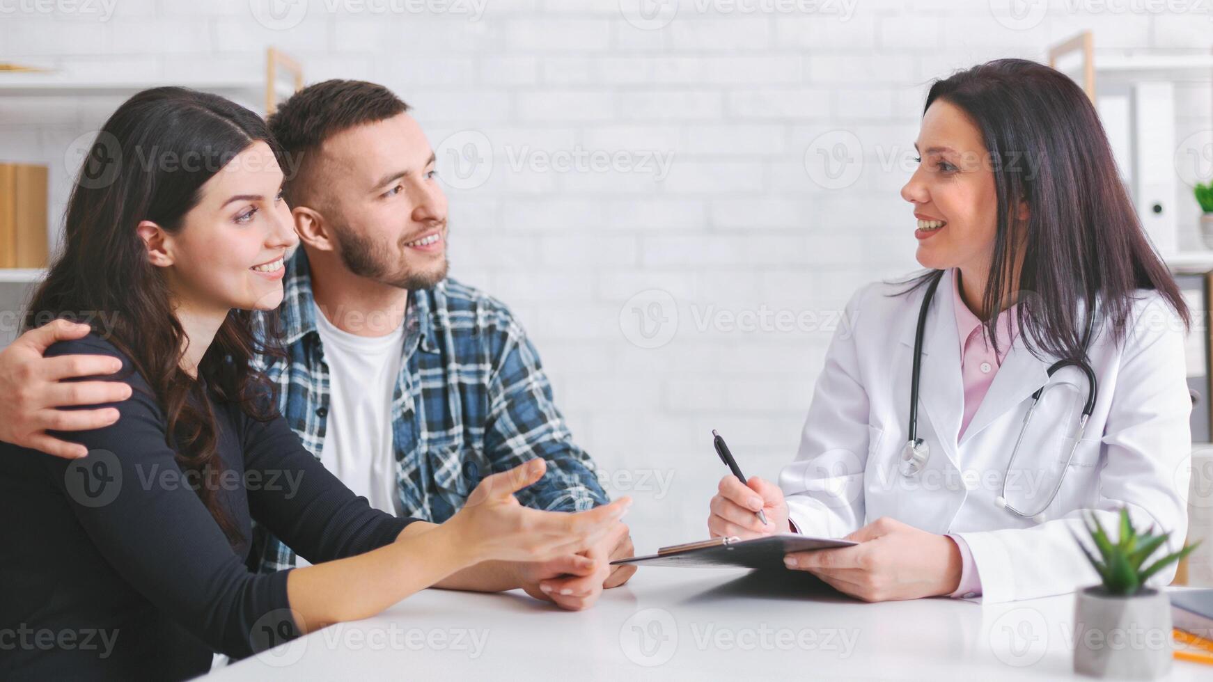European couple sits at a table with woman doctor in a white coat. The couple is engaged in a conversation, listening intently as the doctor takes notes photo
